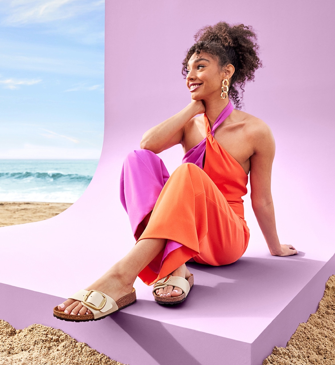 woman in bright purple and orange jumpsuit sitting on beach wearing birkenstock sandals with big buckles